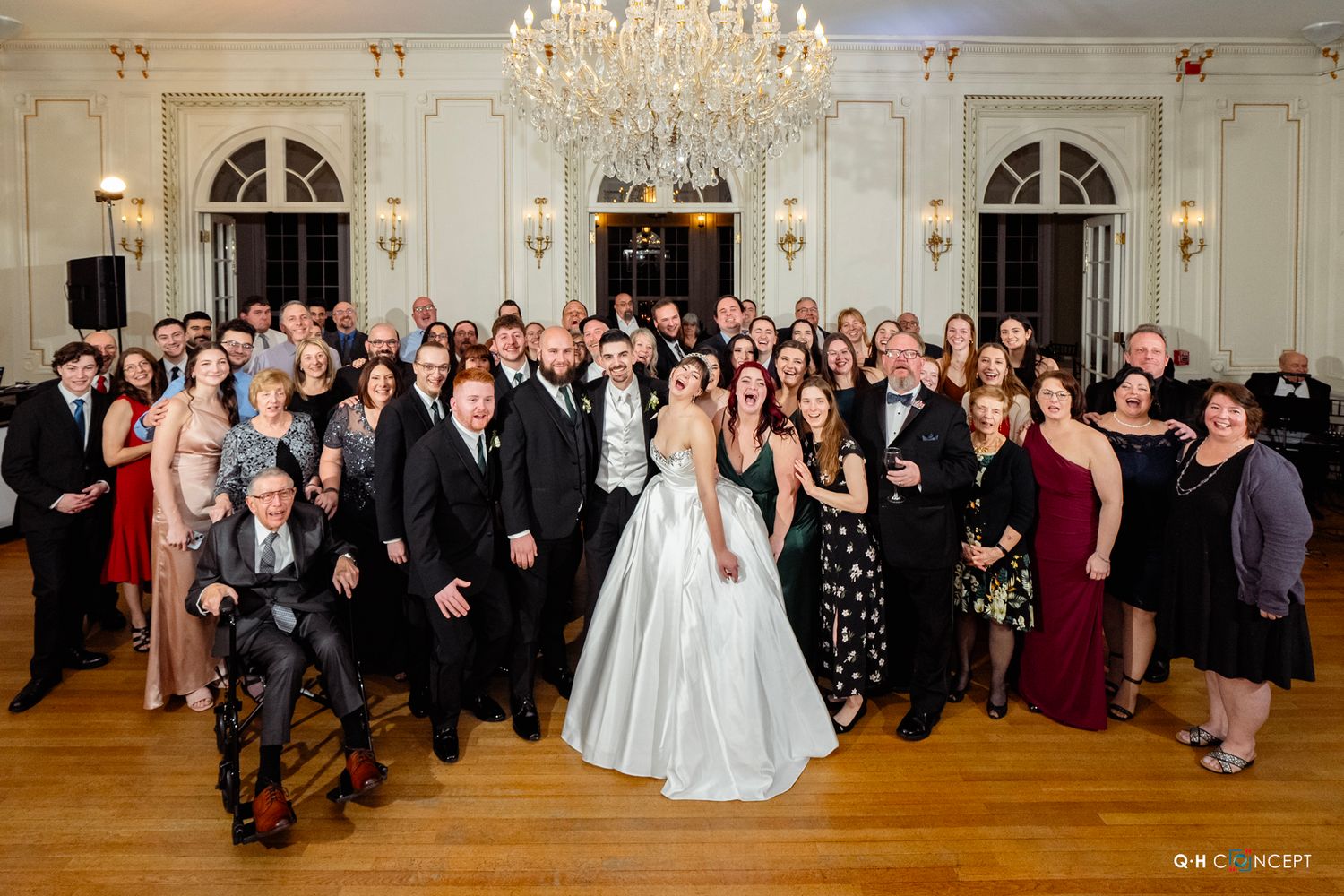 Large group wedding photo with guests in formal attire under an elegant crystal chandelier.