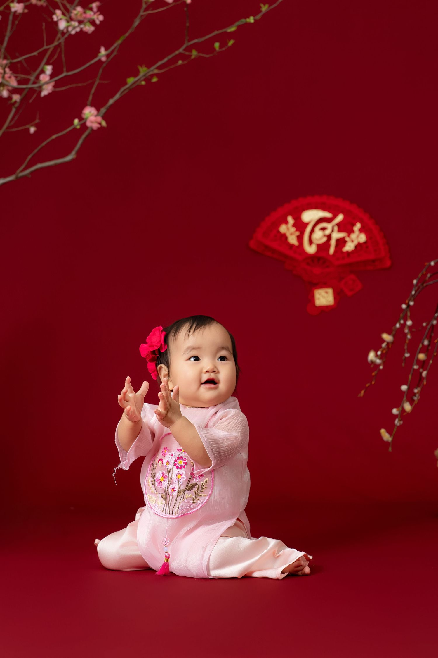 A baby in pink clothing plays with traditional Chinese New Year decorations against a red backdrop with cherry blossoms.