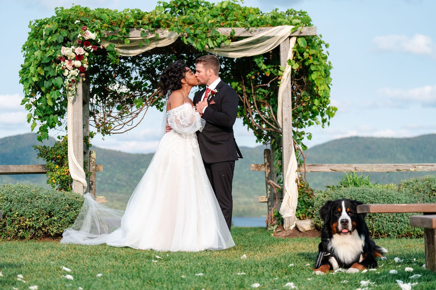 Wedding ceremony under a decorated arch with mountains and lake in the background.