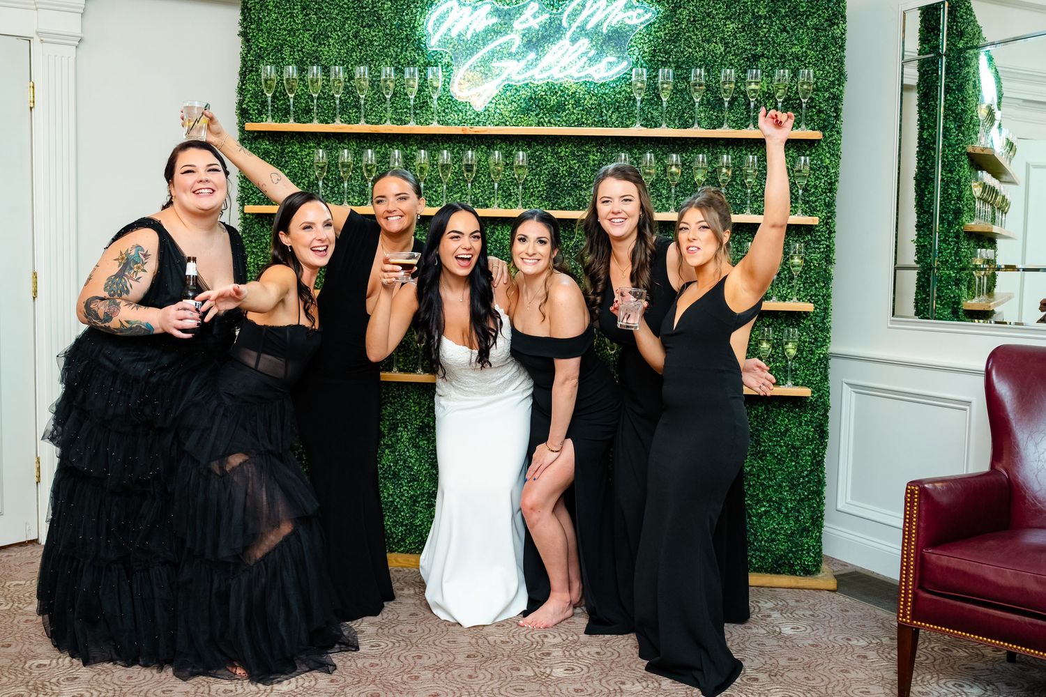 Wedding party in black dresses poses energetically against green wall with champagne display.