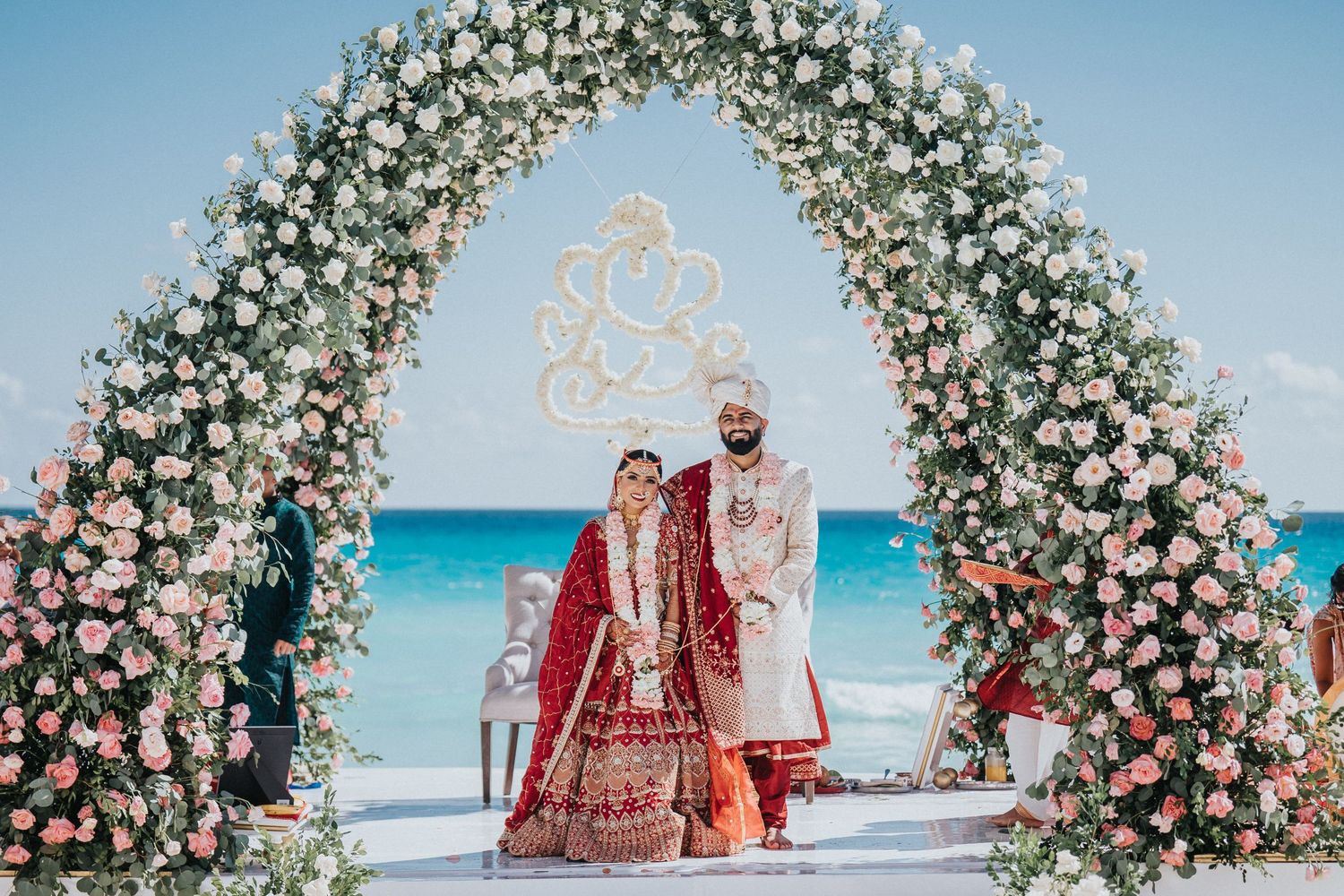 Floral arch decorations frame a traditional Indian wedding ceremony on a scenic beach with turquoise waters.