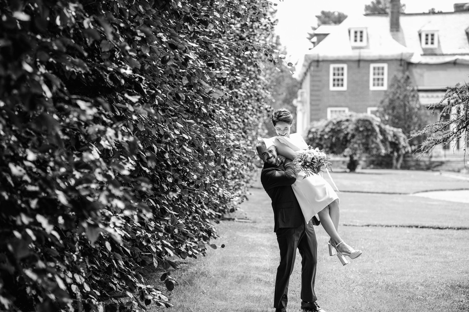 Black and white photo of groom lifting bride in garden near hedge, elegant estate in background.