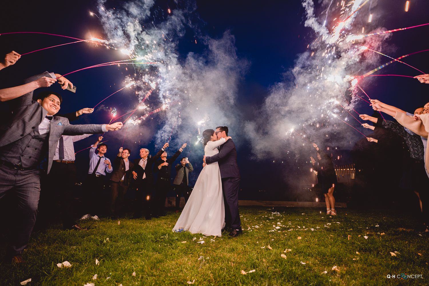 Wedding guests create a sparkler tunnel for the newlyweds on a grassy lawn.