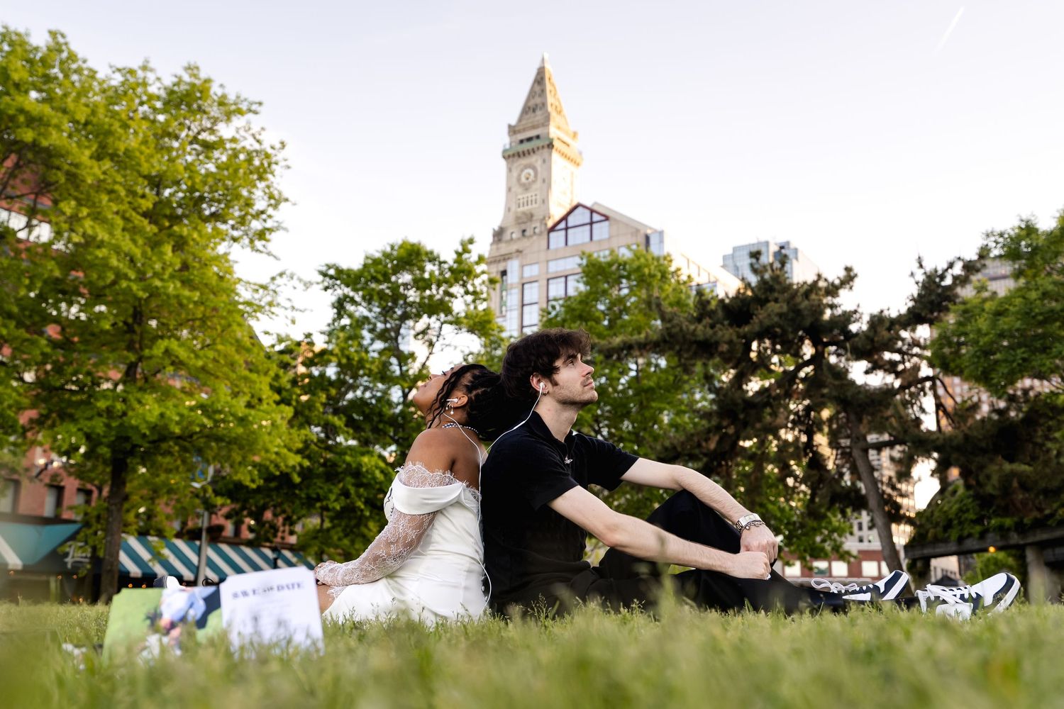 Couple sitting on grass in Boston park with historic Custom House Tower clock visible among trees.