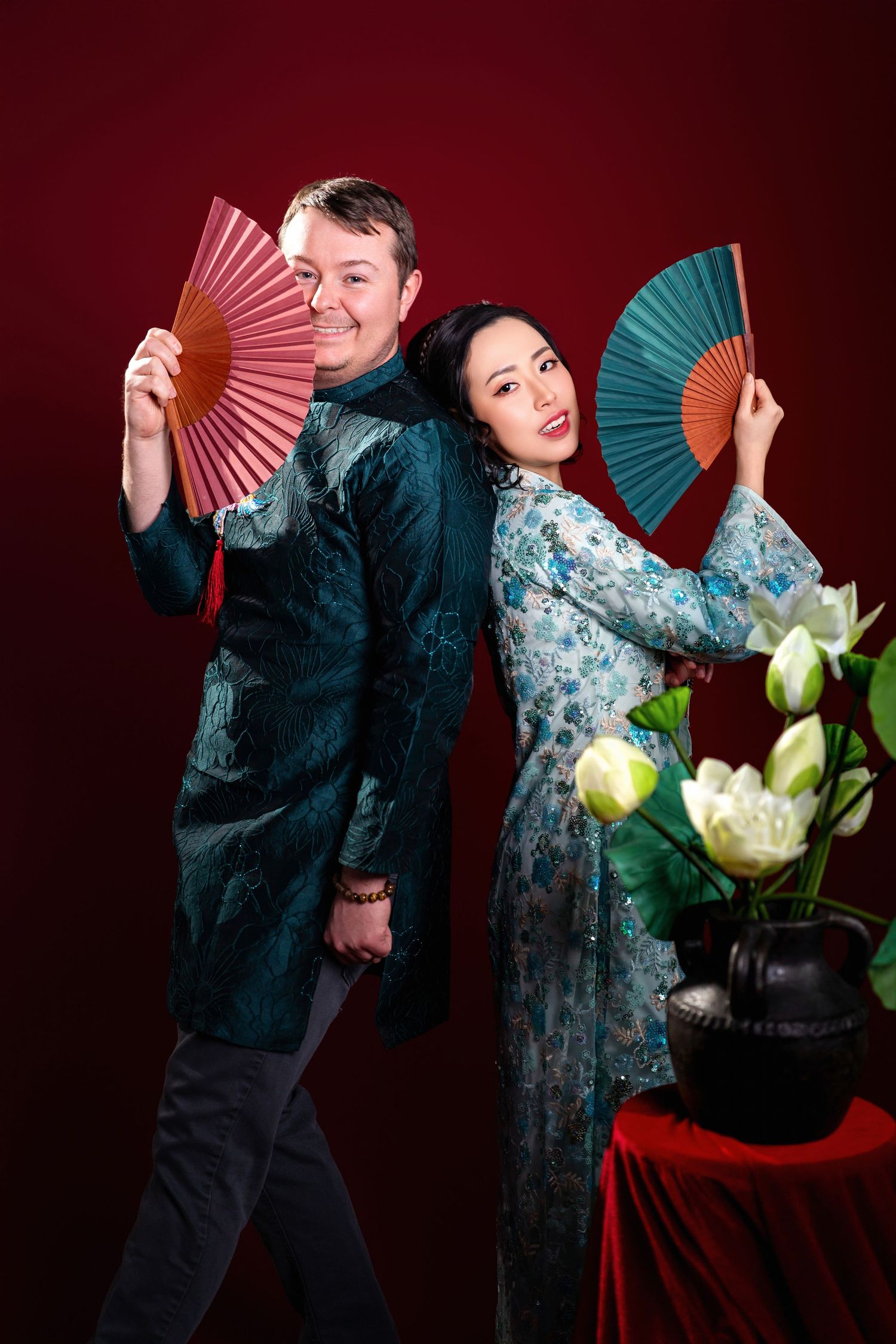 Two performers in traditional Asian costumes pose with decorative fans against a deep red backdrop with white lotus flowers.