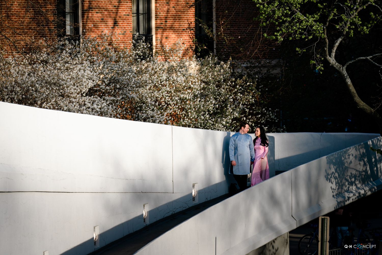 Couple in blue and pink outfits standing on a modern white bridge surrounded by blooming spring trees.