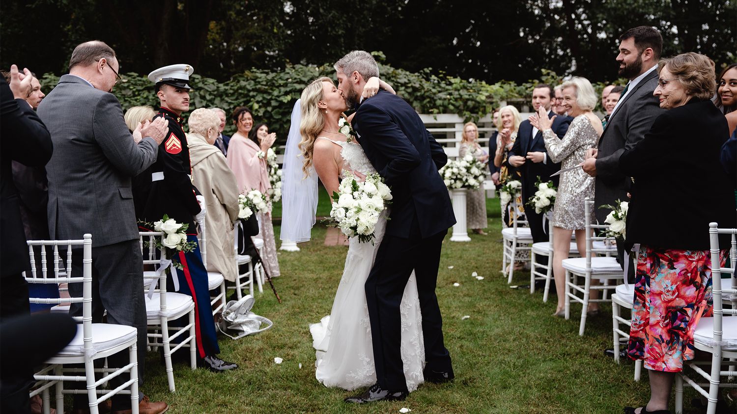 Panoramic view of an outdoor wedding ceremony with white chairs on a green lawn surrounded by trees and guests.