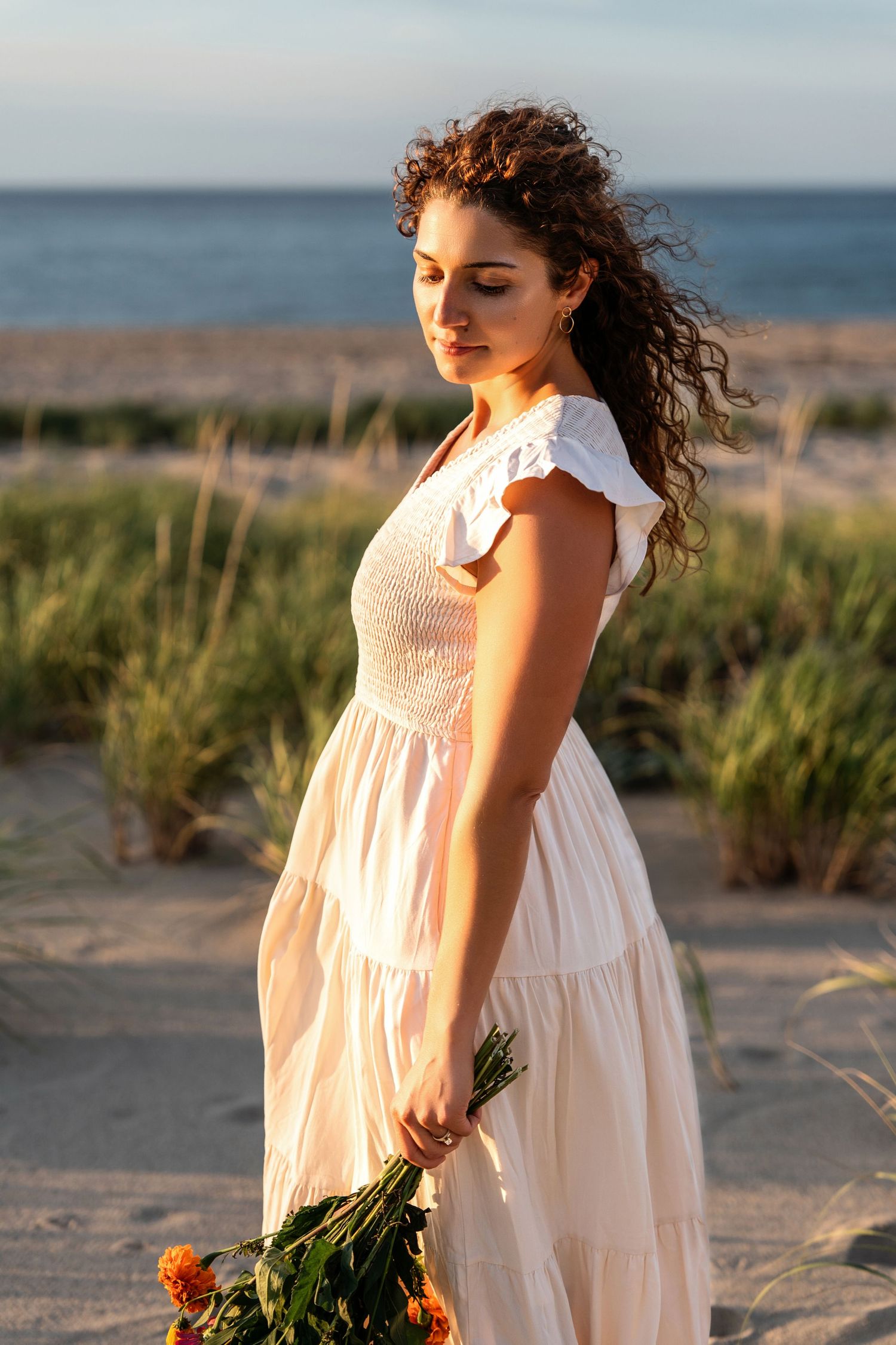 Person in white dress holding flowers on beach at golden hour.
