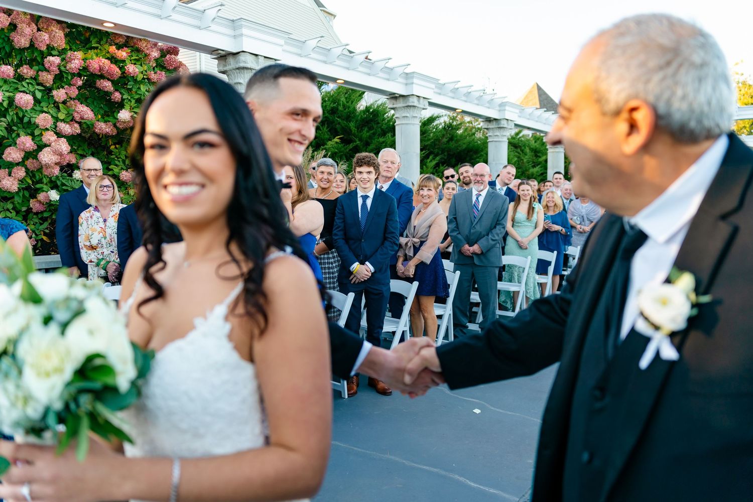A wedding ceremony takes place under a white pergola with pink flowers and gathered guests.