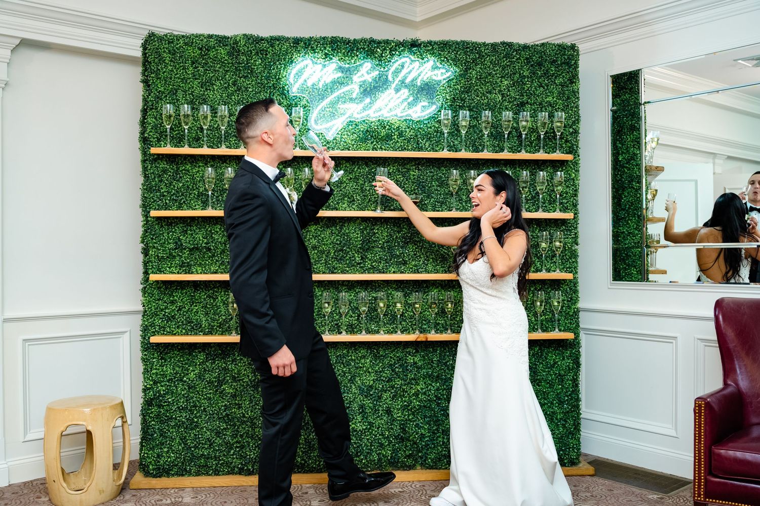 Wedding couple poses against a lush green hedge wall with champagne flutes.