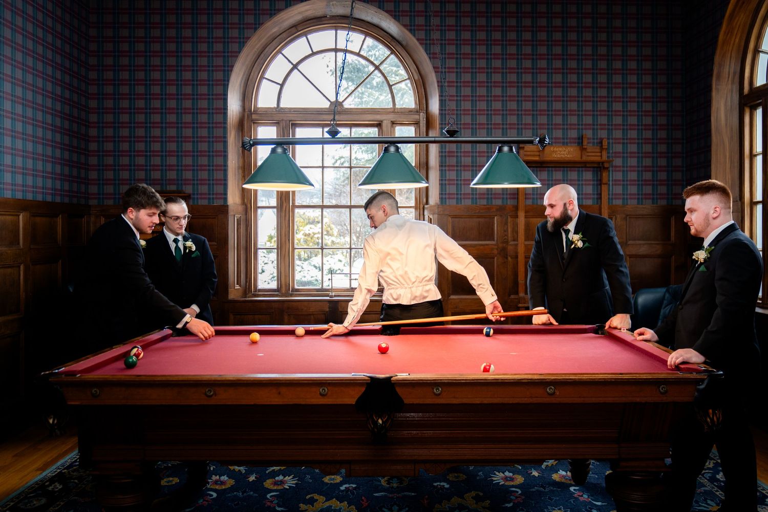 People playing pool at a billiards table in an elegant room with arched windows and pendant lighting.