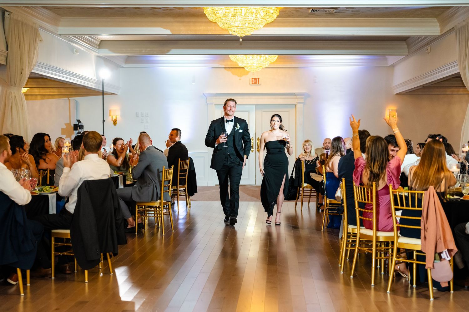 Wide angle shot of wedding party dancing in a luxurious white ballroom with crystal chandeliers.
