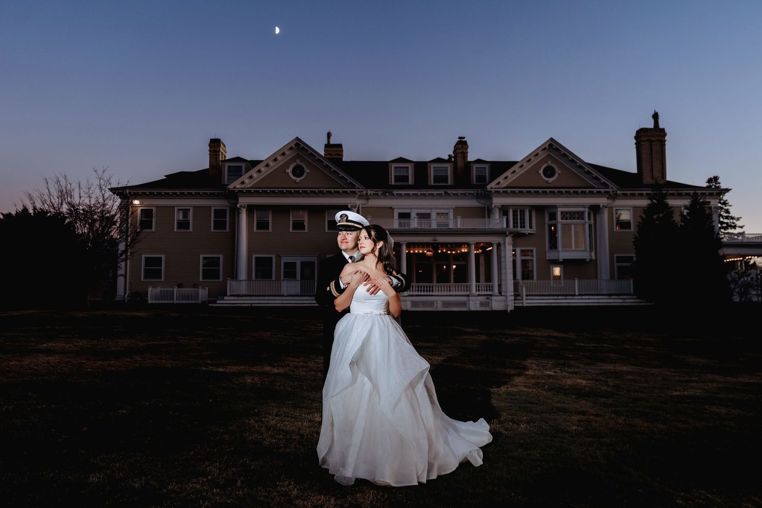 A bride in a flowing white gown twirls on a lawn at sunset with a grand estate building in the background.