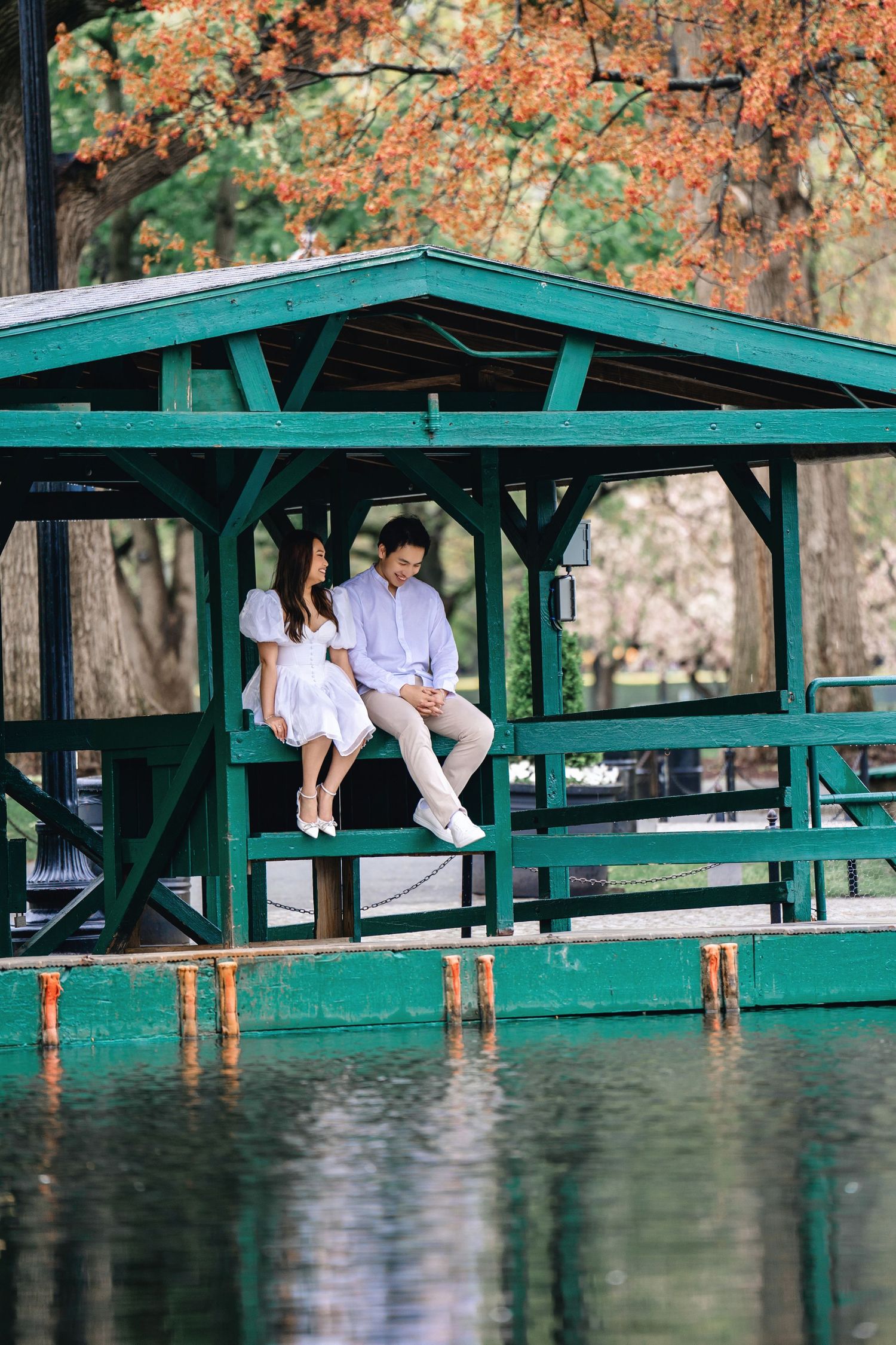 People sitting on green wooden picnic tables under pavilion with autumn trees and reflective water in background.