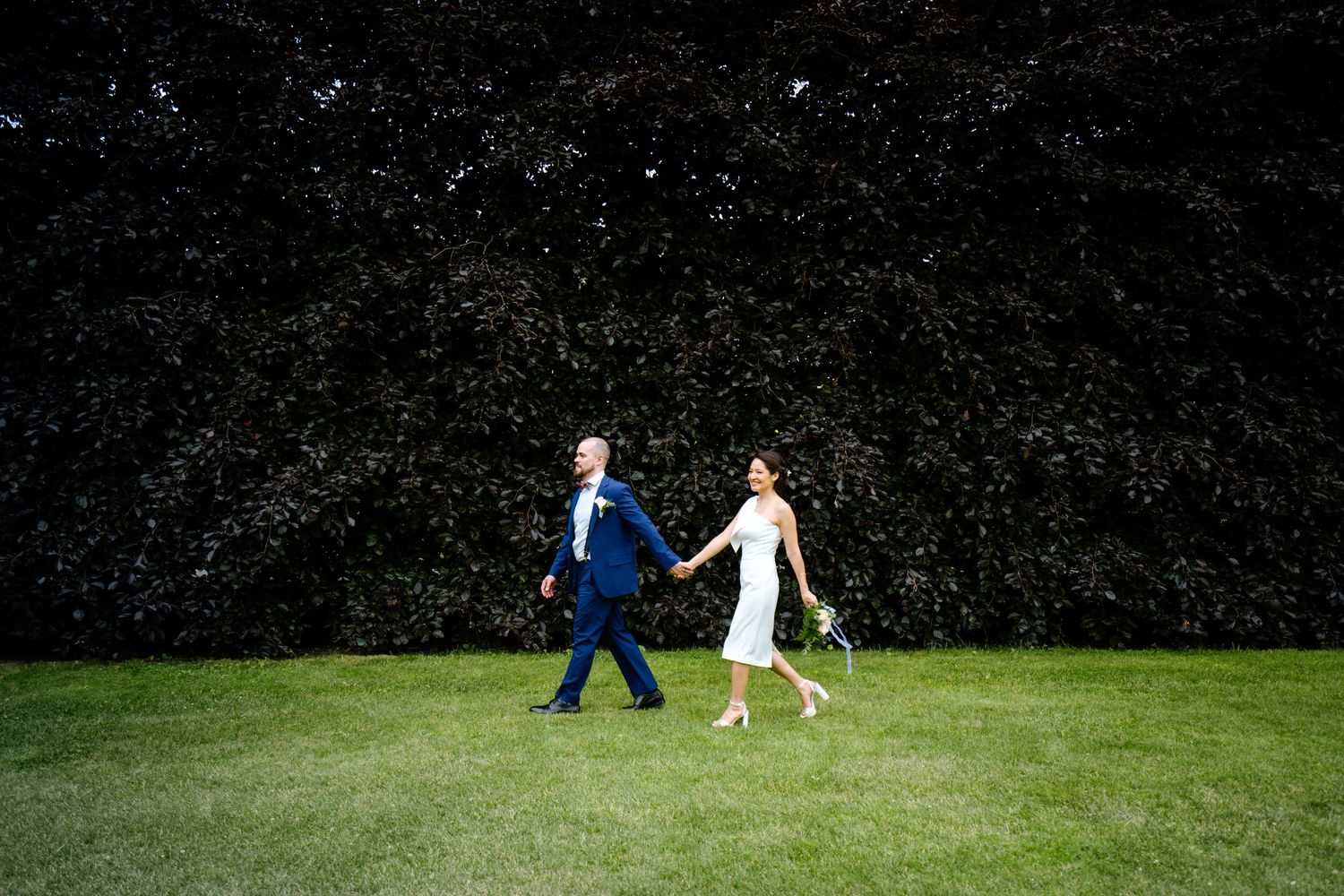 A bride and groom walk hand in hand across a lush green lawn in front of a dark ivy-covered wall.