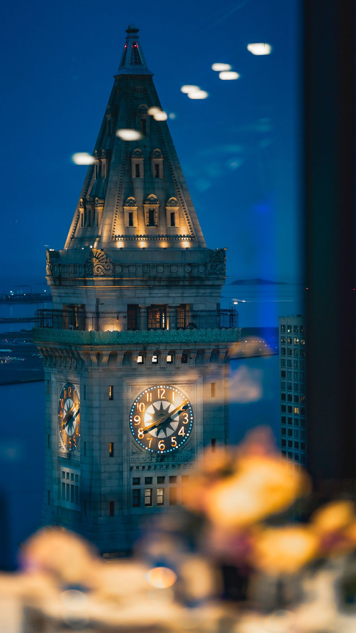 Illuminated clock tower of Boston Custom House glows against dark blue evening sky with blurred foreground elements.