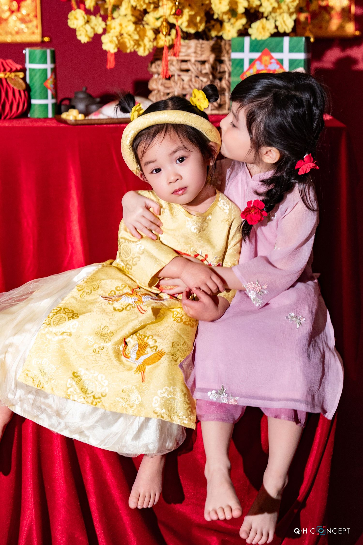 Two children in yellow and pink traditional dresses sit playfully on red fabric under golden decorations.