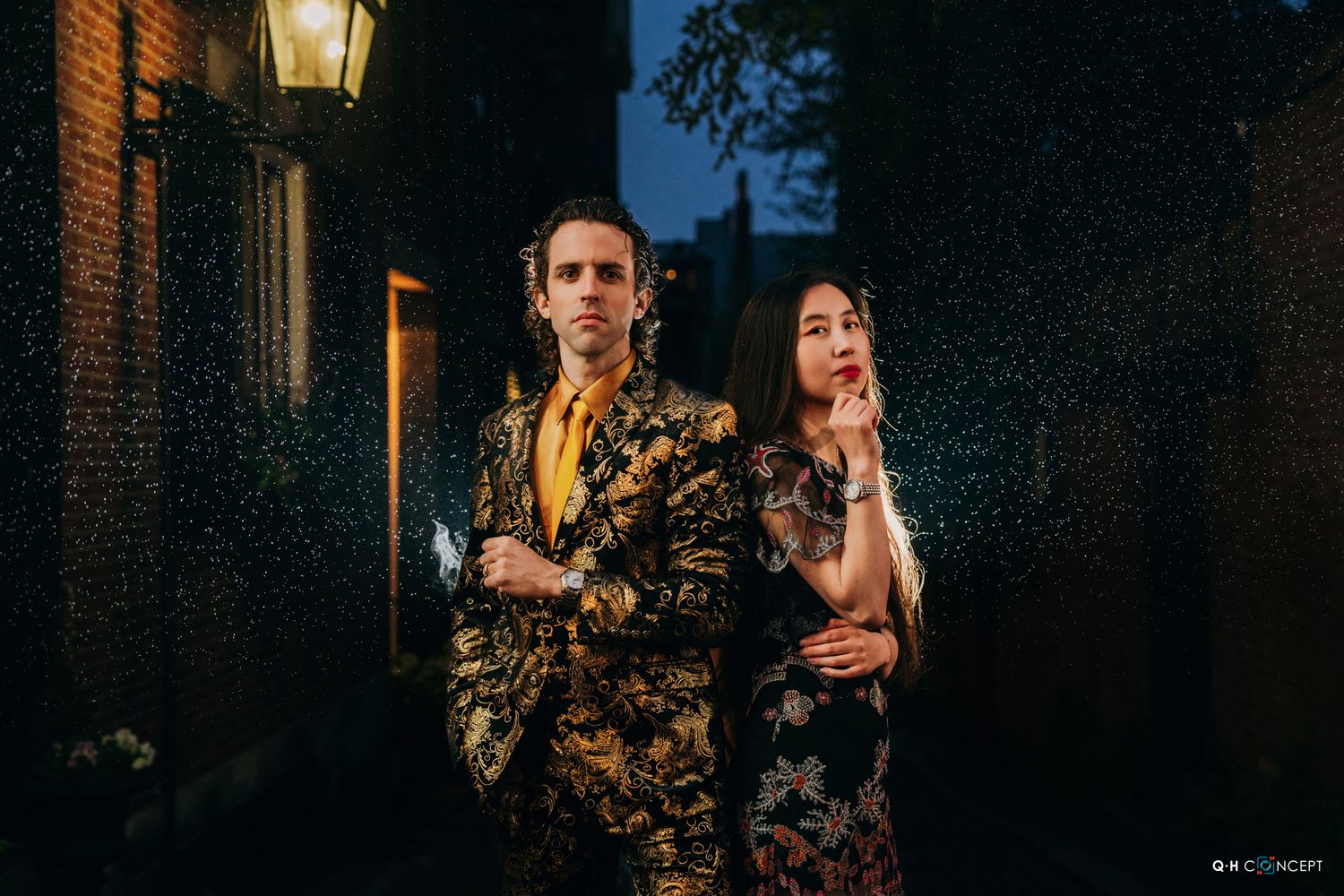 Couple shares a moment under vintage street lamp against brick wall at night.