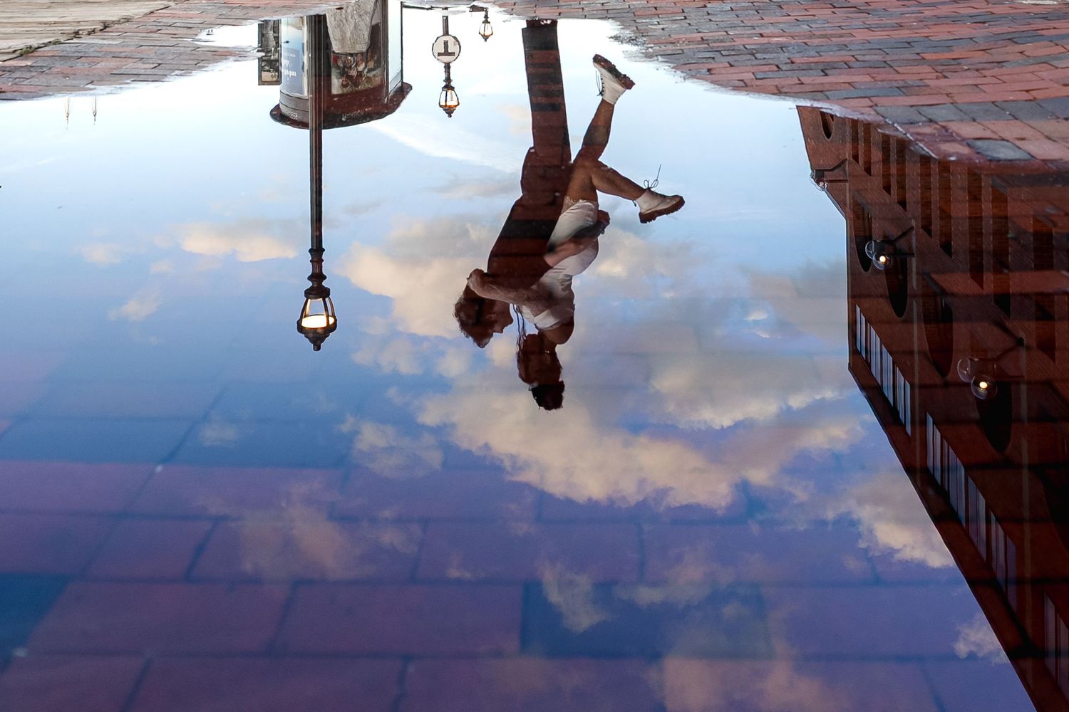 Person reflected upside down in a rain puddle on brick pavement with clouds and buildings mirrored.