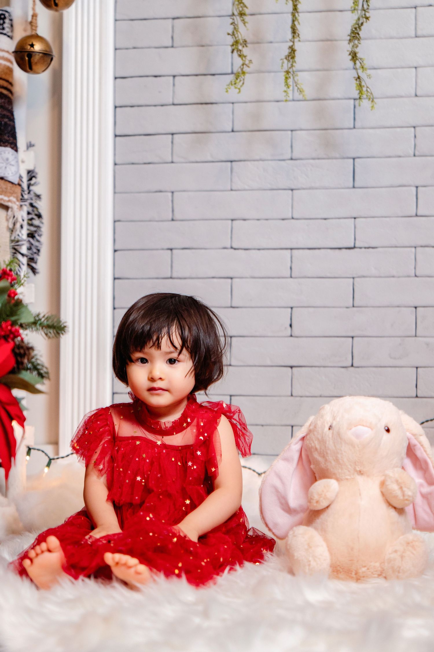 A child in a sparkly red dress poses on white faux fur with a pink plush toy in a holiday-themed studio setting.