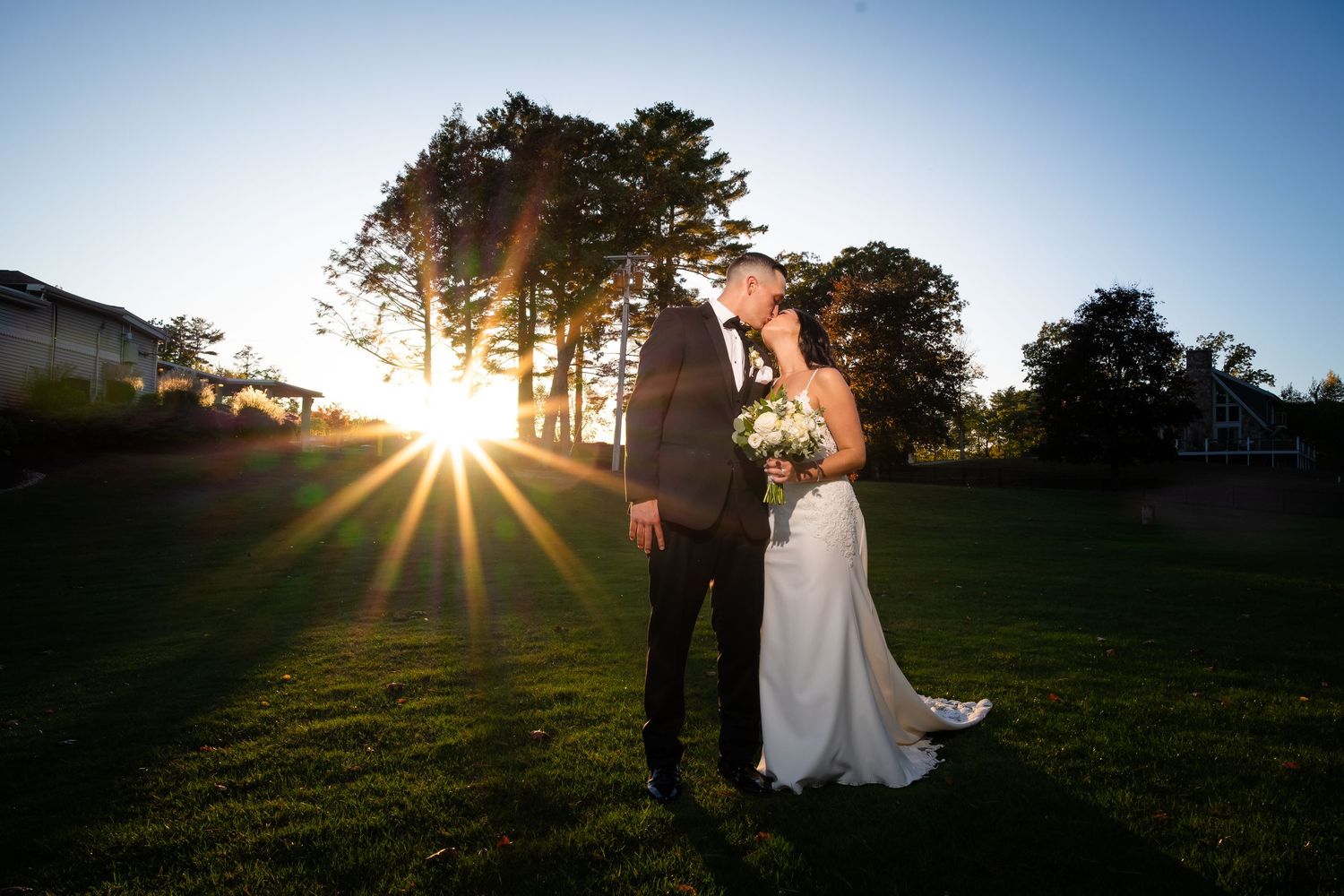 Dramatic sunburst peeks through trees silhouetting figures against a bright blue sky at dusk.