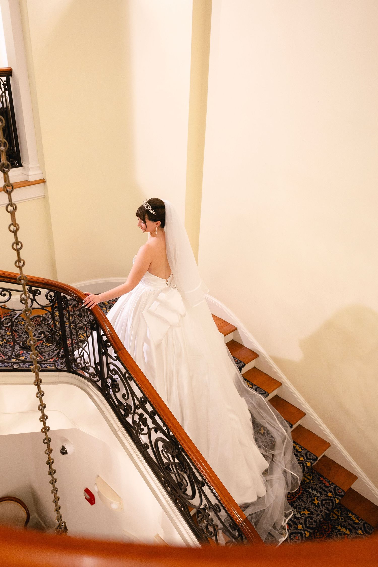 A bride in a flowing white wedding dress descends an ornate staircase with dark wrought iron railings.