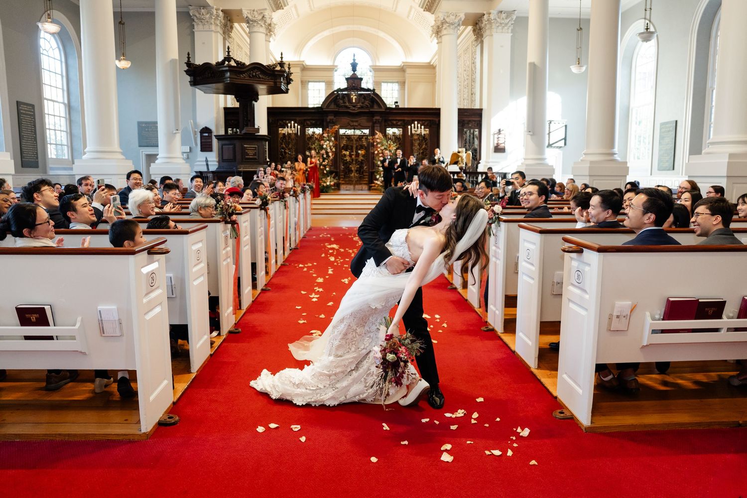 A bride and groom walk down a red carpeted church aisle while guests throw white flower petals in celebration.