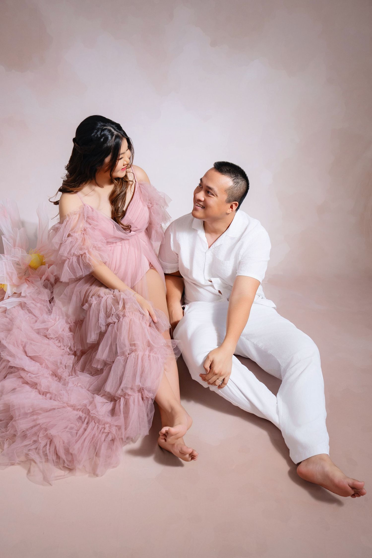 Romantic maternity session with couple in pink tulle dress laying on light colored floor.