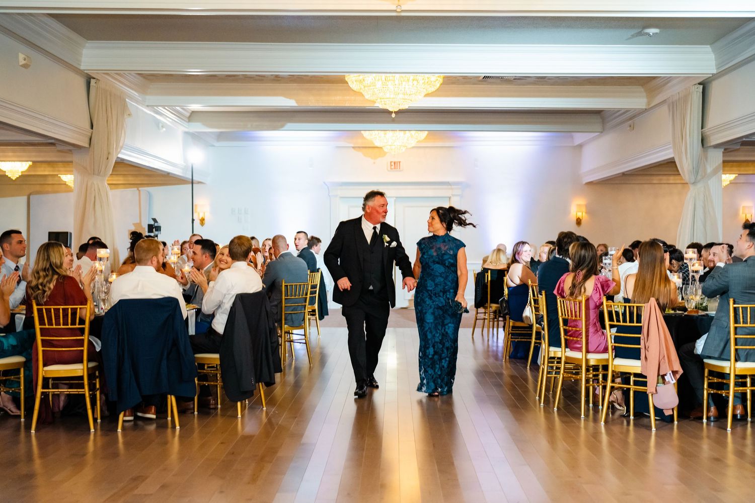 Wedding reception in a bright ballroom with guests seated at round tables and chandeliers overhead.