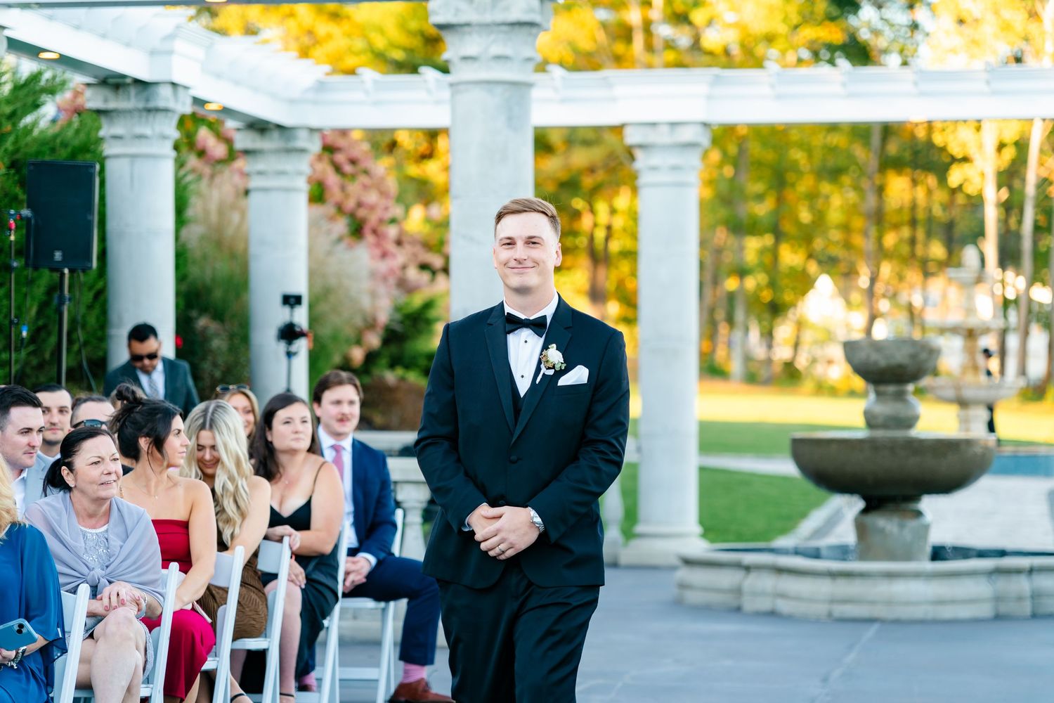 Groom stands at altar under white columned pergola during outdoor autumn ceremony.