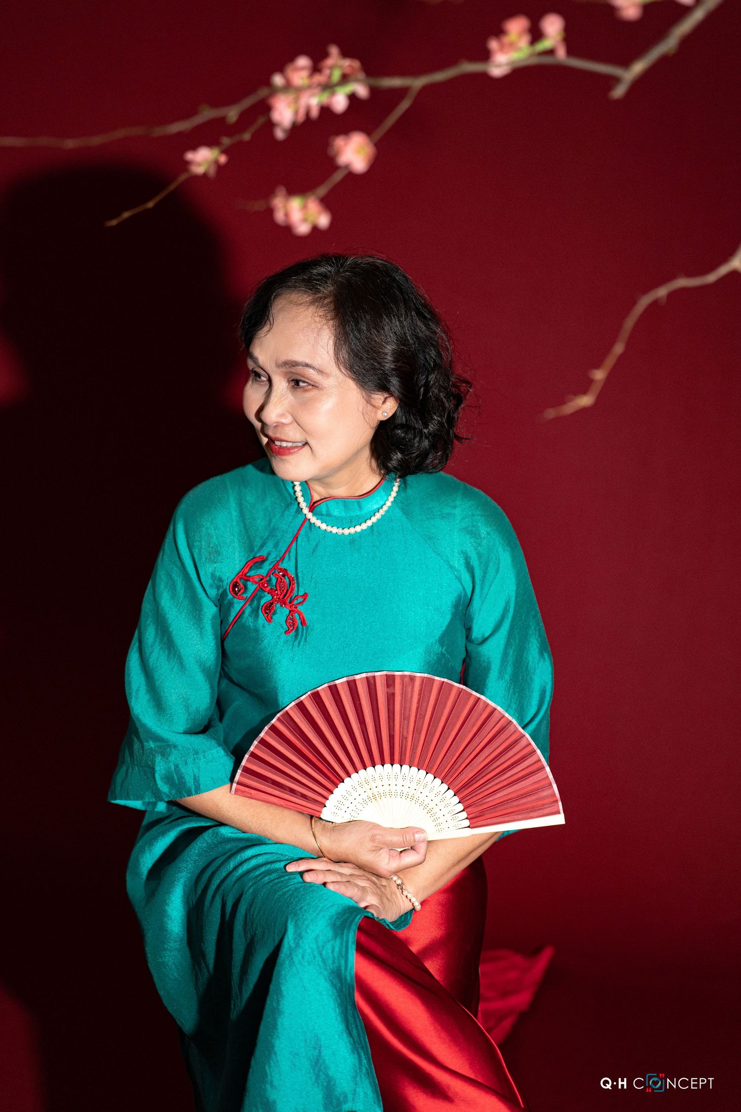 Woman in turquoise traditional dress holding red folding fan against burgundy backdrop with cherry blossoms.