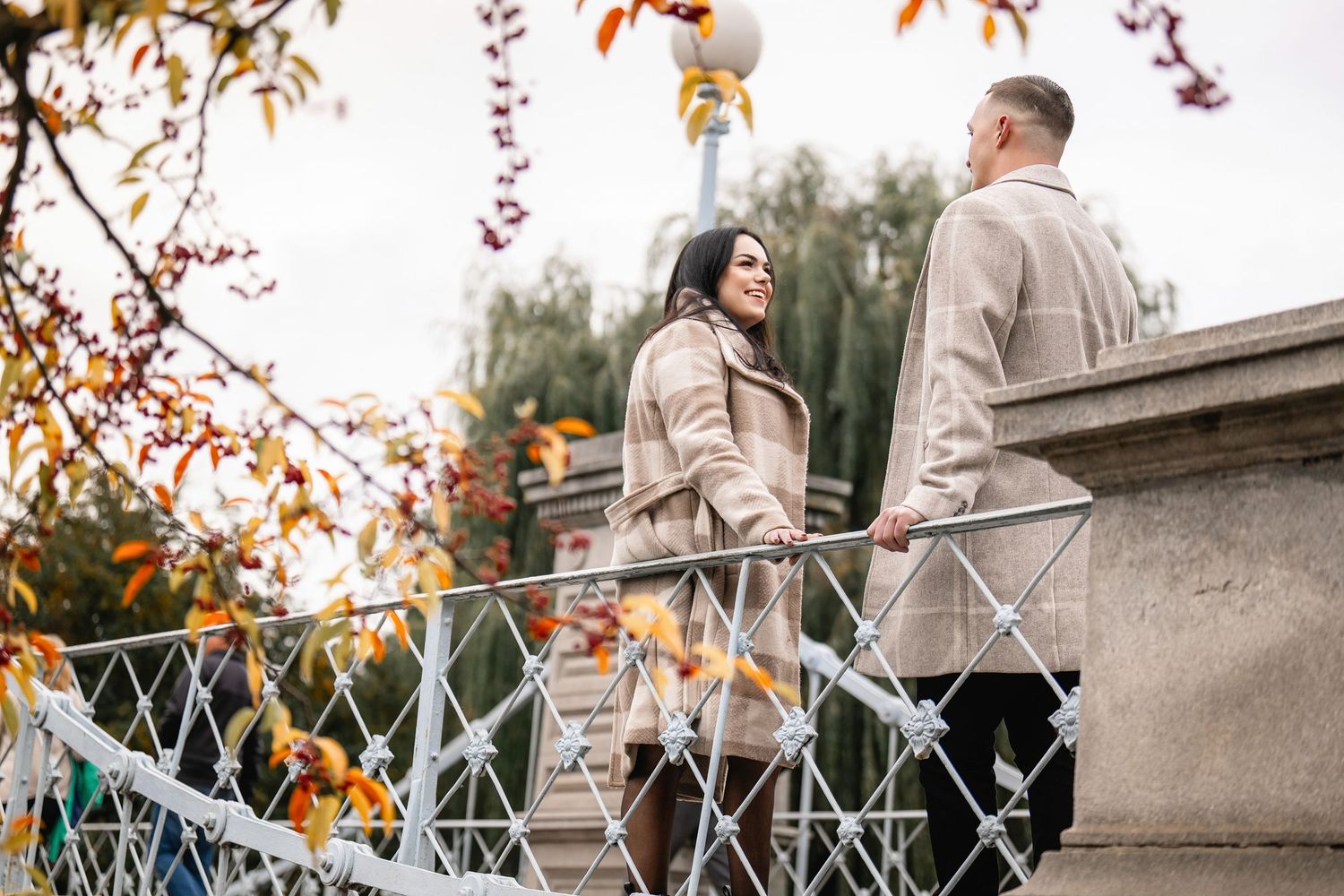 Couple walking together along a stone bridge railing with autumn leaves in the background during a fall photoshoot.
