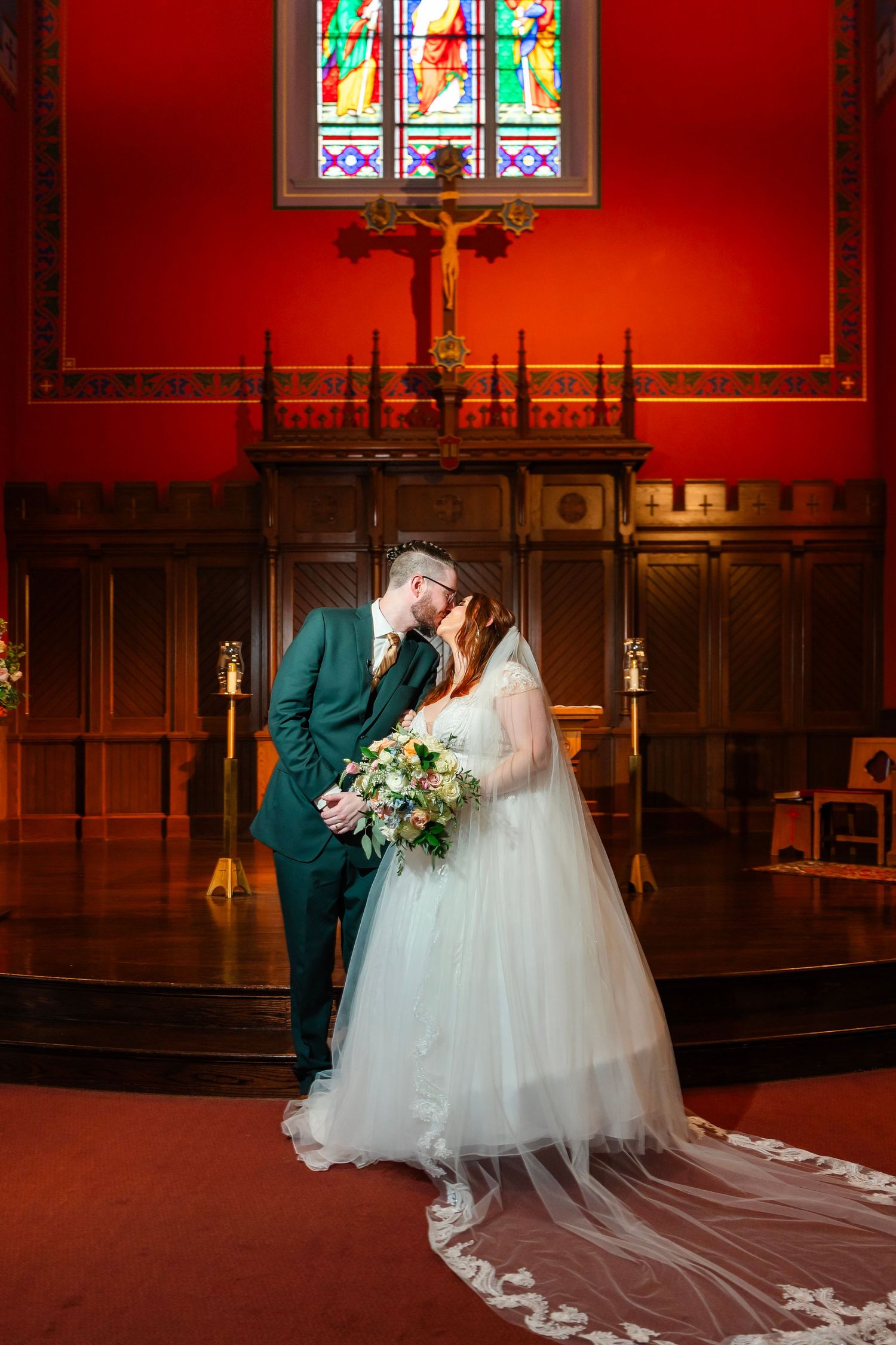 A romantic wedding ceremony in a church sanctuary with red walls and stained glass windows.