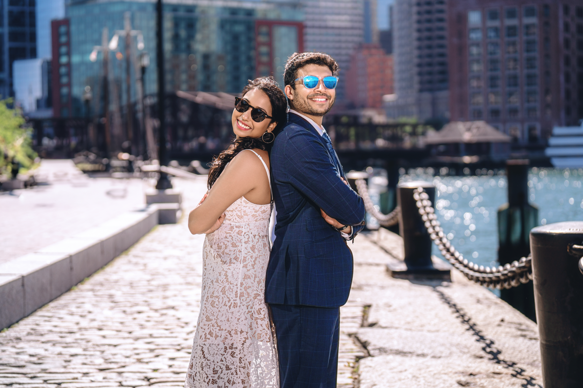 A couple shares an intimate moment along a waterfront with urban buildings in the background.