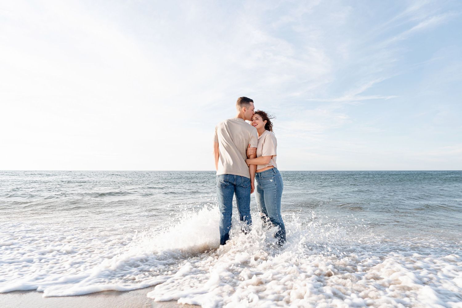 A couple stands romantically at the ocean's edge, waves splashing around their feet on a sunny beach day.