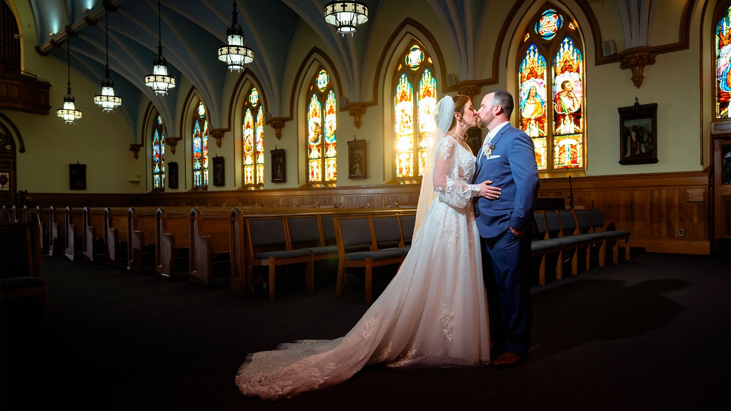 A couple stands together in a beautiful gothic church with stained glass windows and ornate wooden pews.