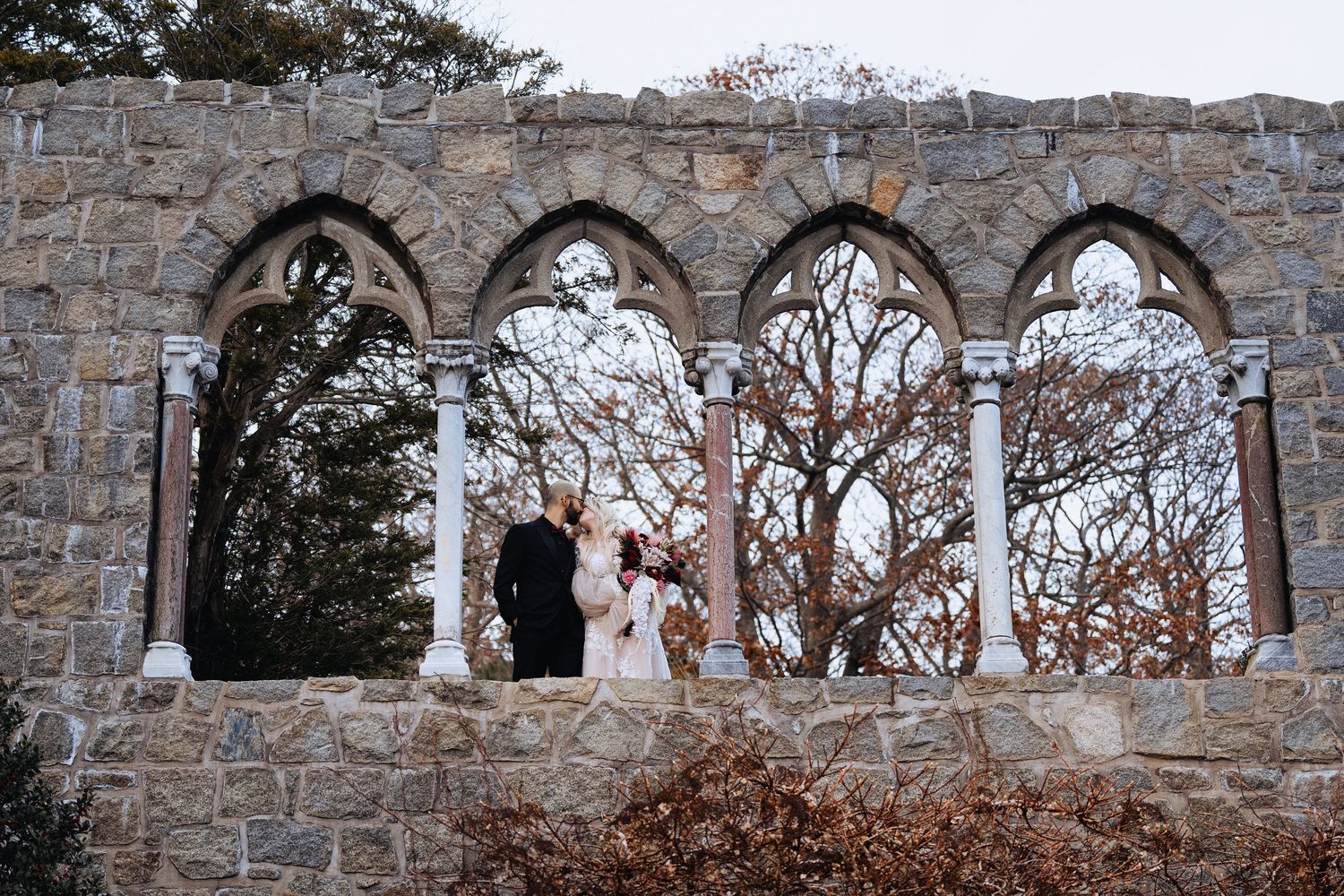 Stone arches of a historic building frame a scenic autumn landscape with bare trees and cloudy skies.