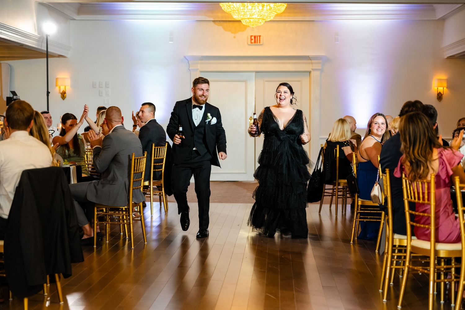 Wedding guests dancing and celebrating in an elegant ballroom with chandeliers and gold chairs.