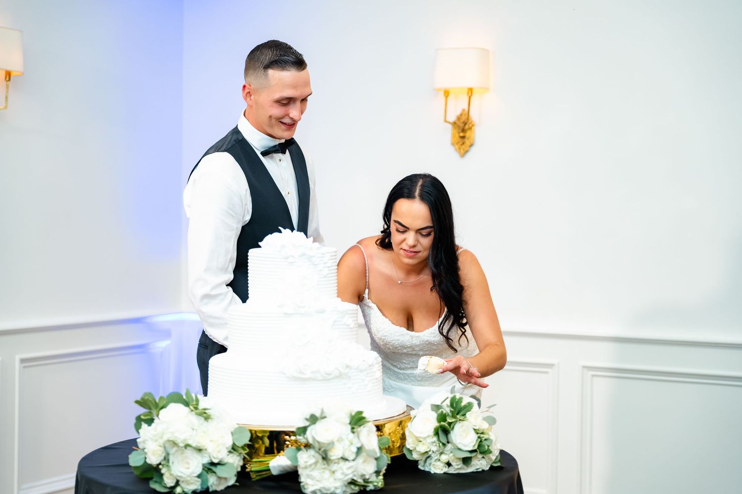 Wedding party enjoying first dances in an elegant white reception hall with floral centerpieces.