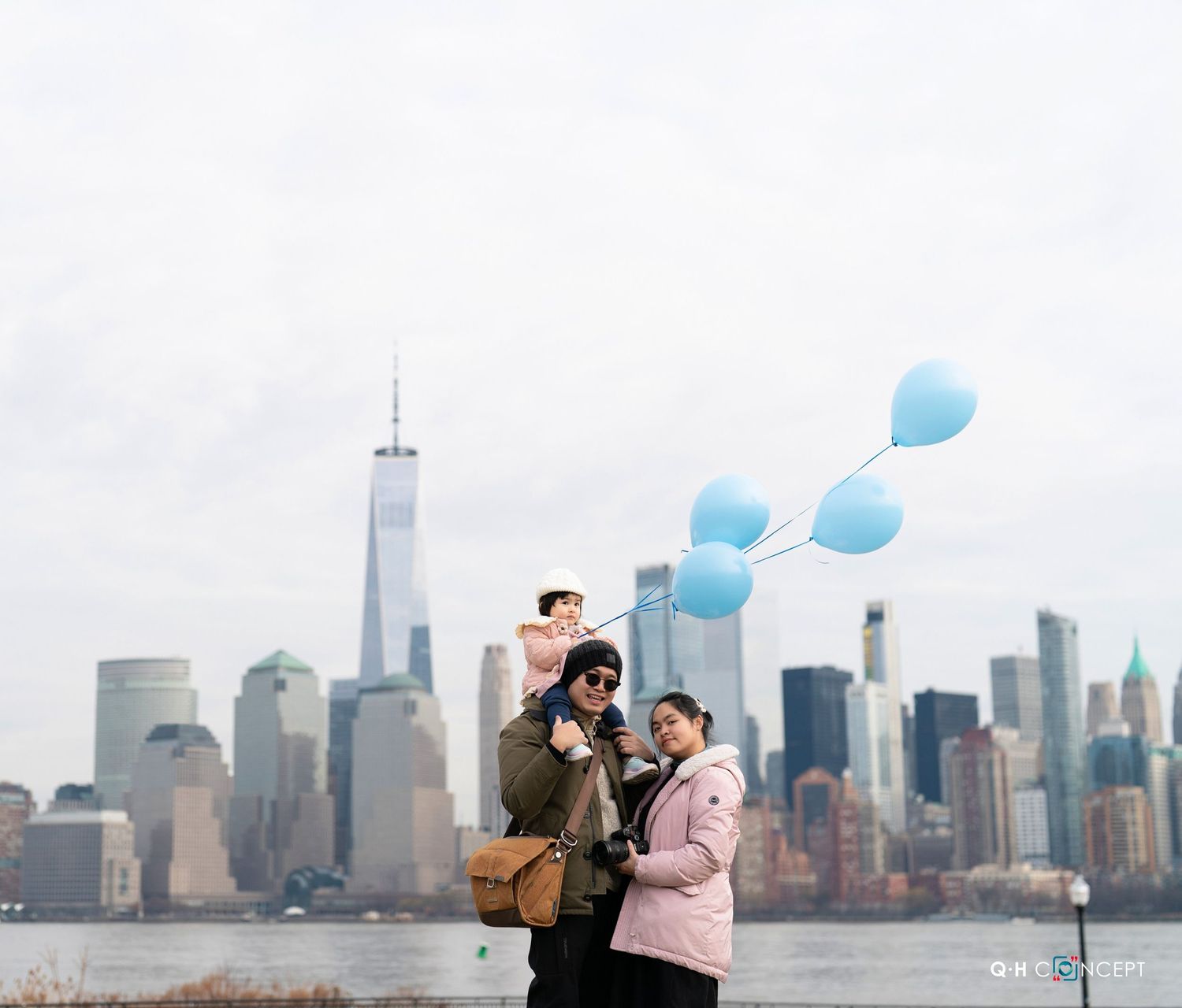 Family with blue balloons posing by the Hudson River with the NYC skyline and One World Trade Center in the background.