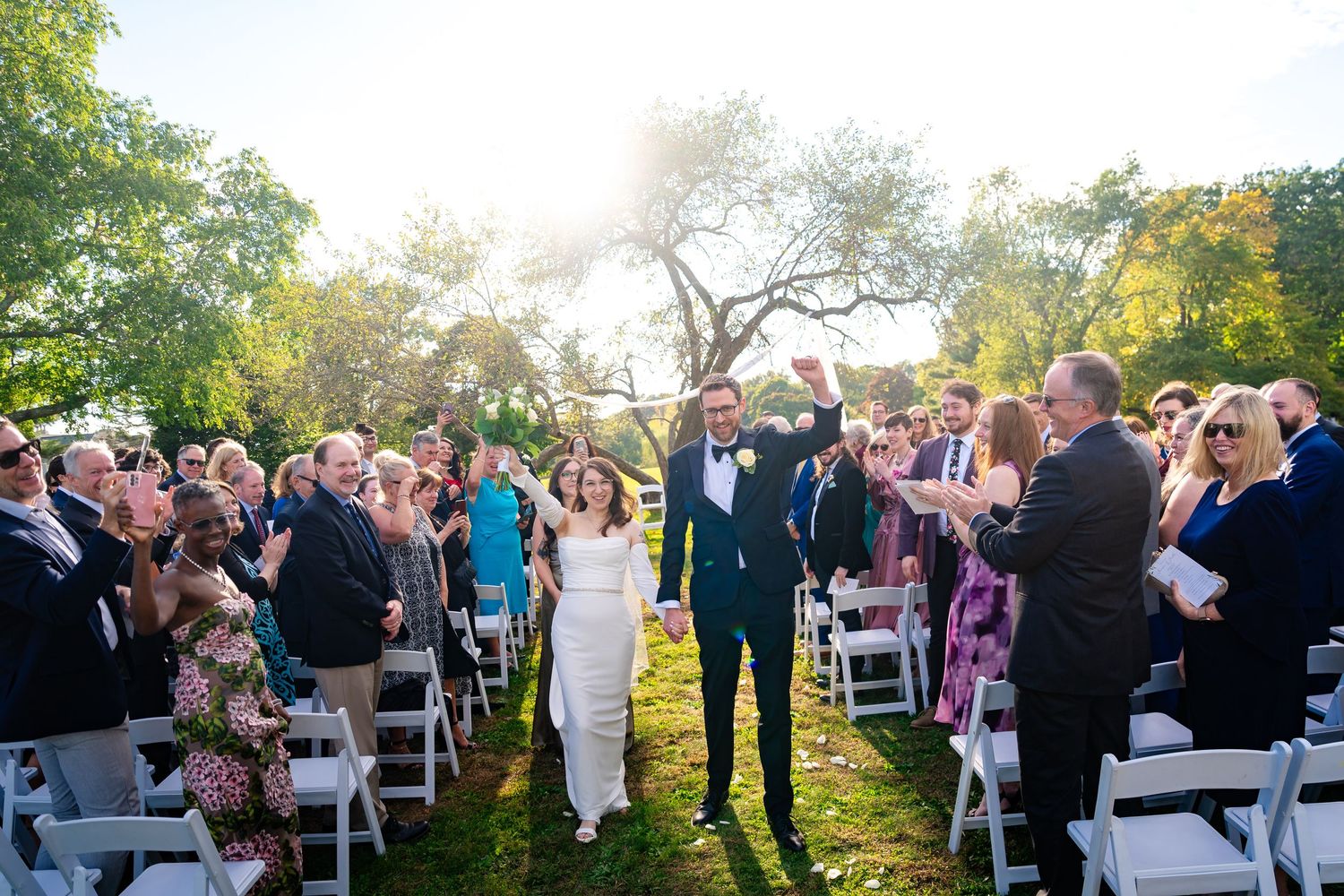 Wedding guests mingle during an outdoor ceremony with trees and golden sunlight.