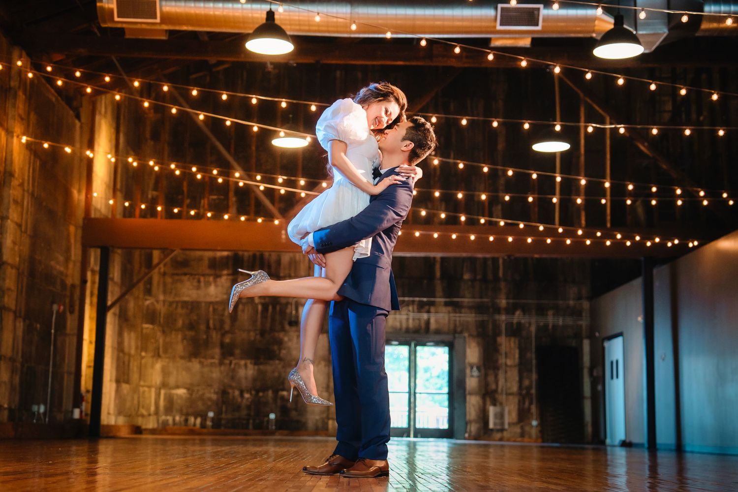 Wedding couple dances in rustic venue decorated with overhead string lights.