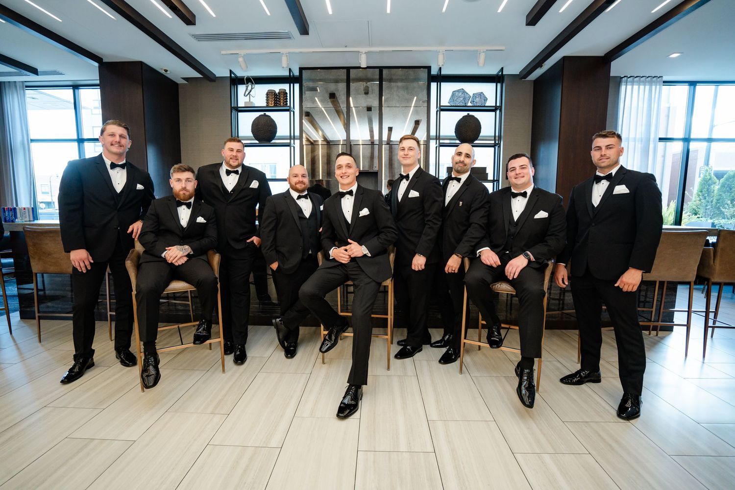 Groomsmen in formal attire pose sitting in modern venue with floor to ceiling windows.