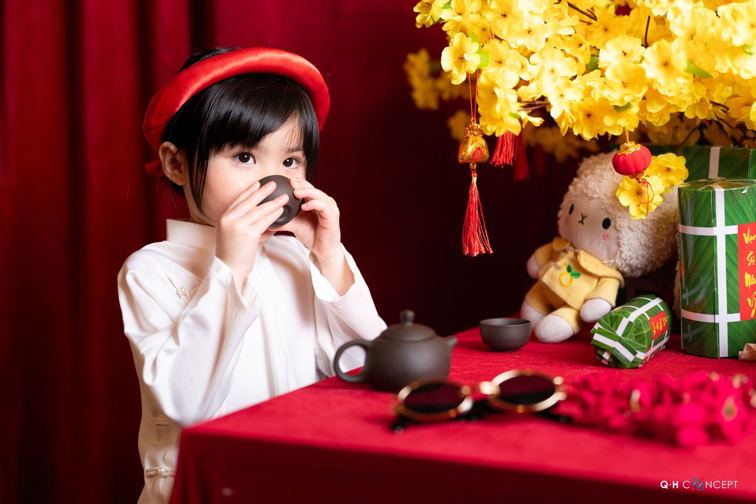 A child in a white shirt sits near a Christmas tree and holiday decorations with a red backdrop.