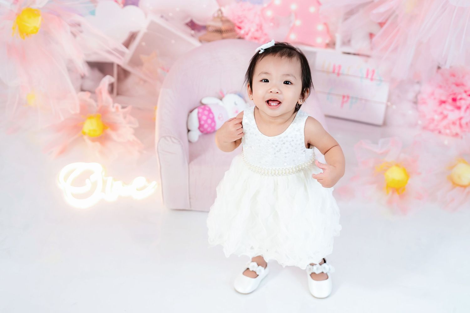 A small child in a white dress smiles while standing against a pink floral backdrop.
