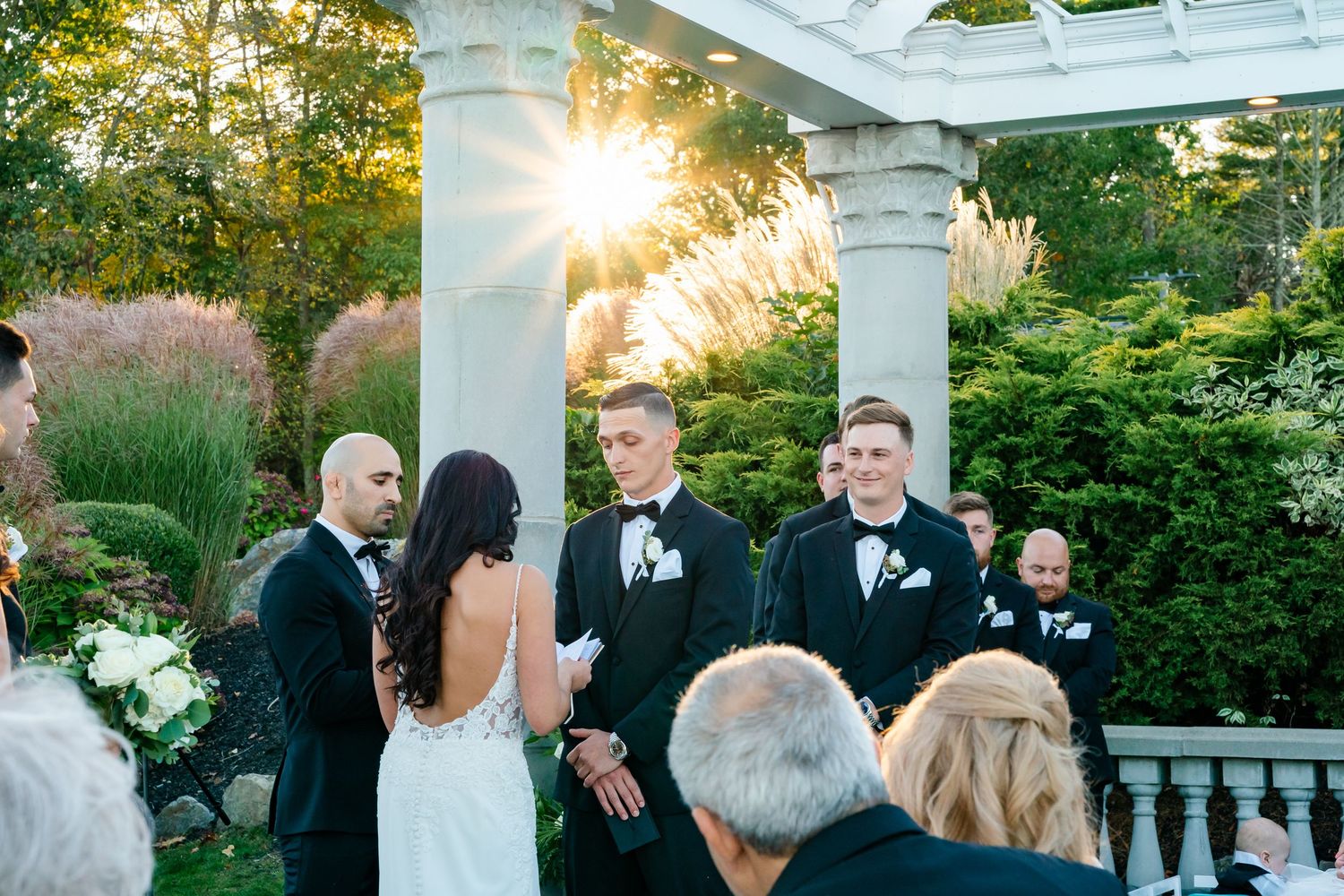 Wedding guests gather outdoors at sunset in a garden setting with elegant formal attire.
