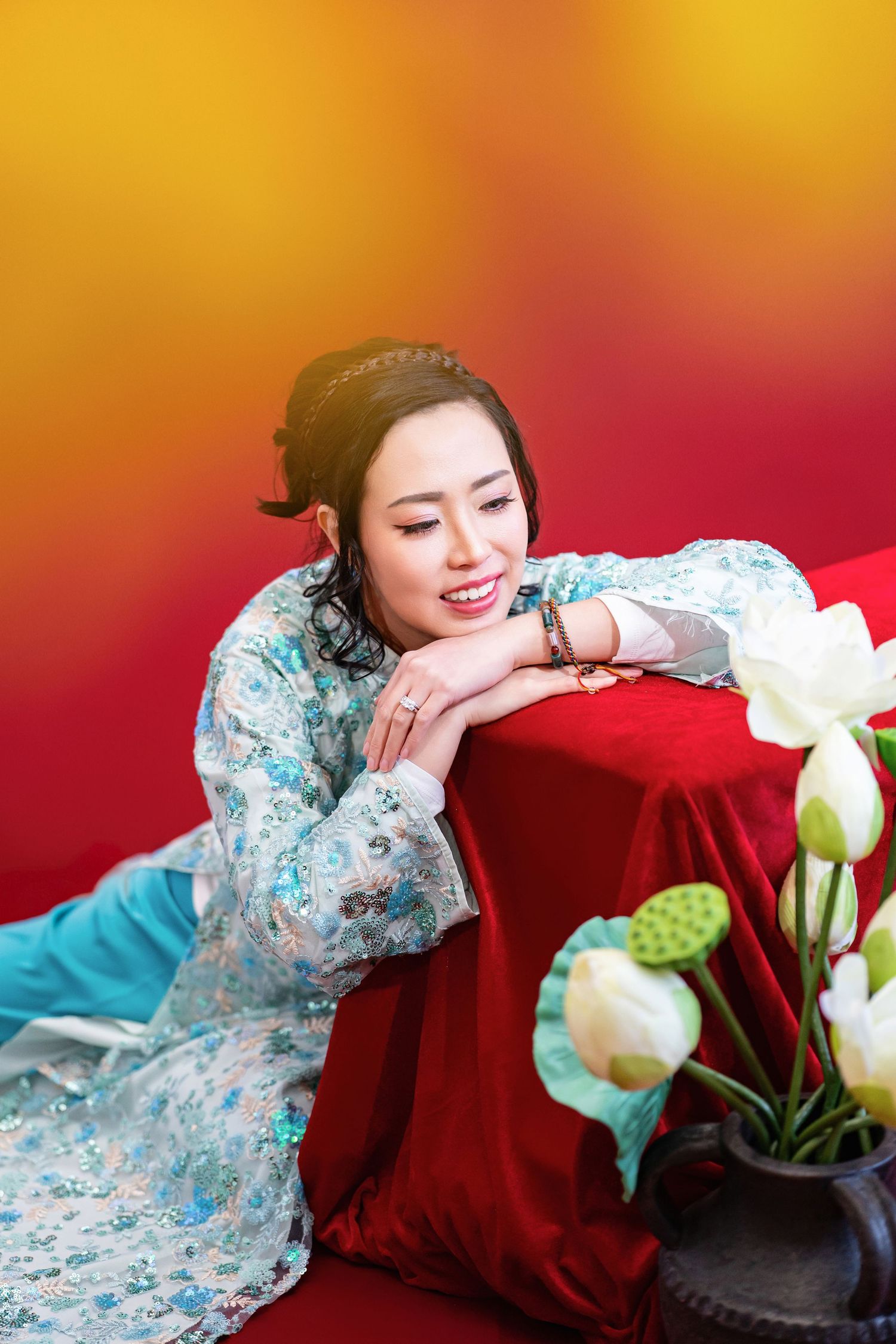 A person in a blue floral dress rests on a red velvet couch next to white flowers in a vase.