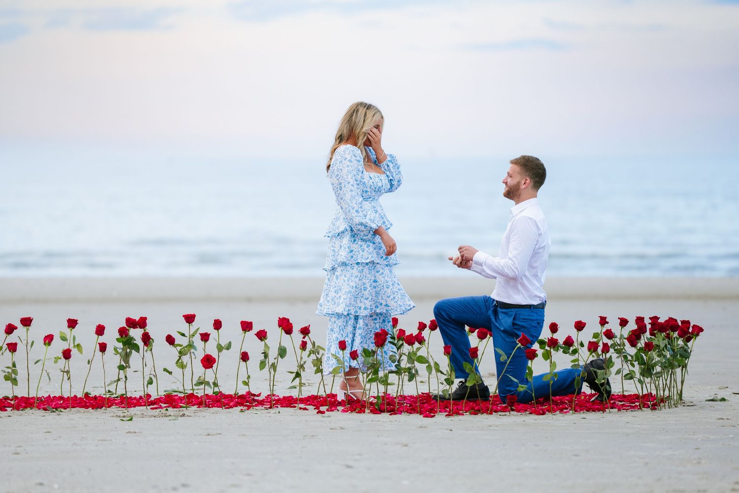 Romantic beach proposal scene with red rose petals arranged in a heart shape on white sand.