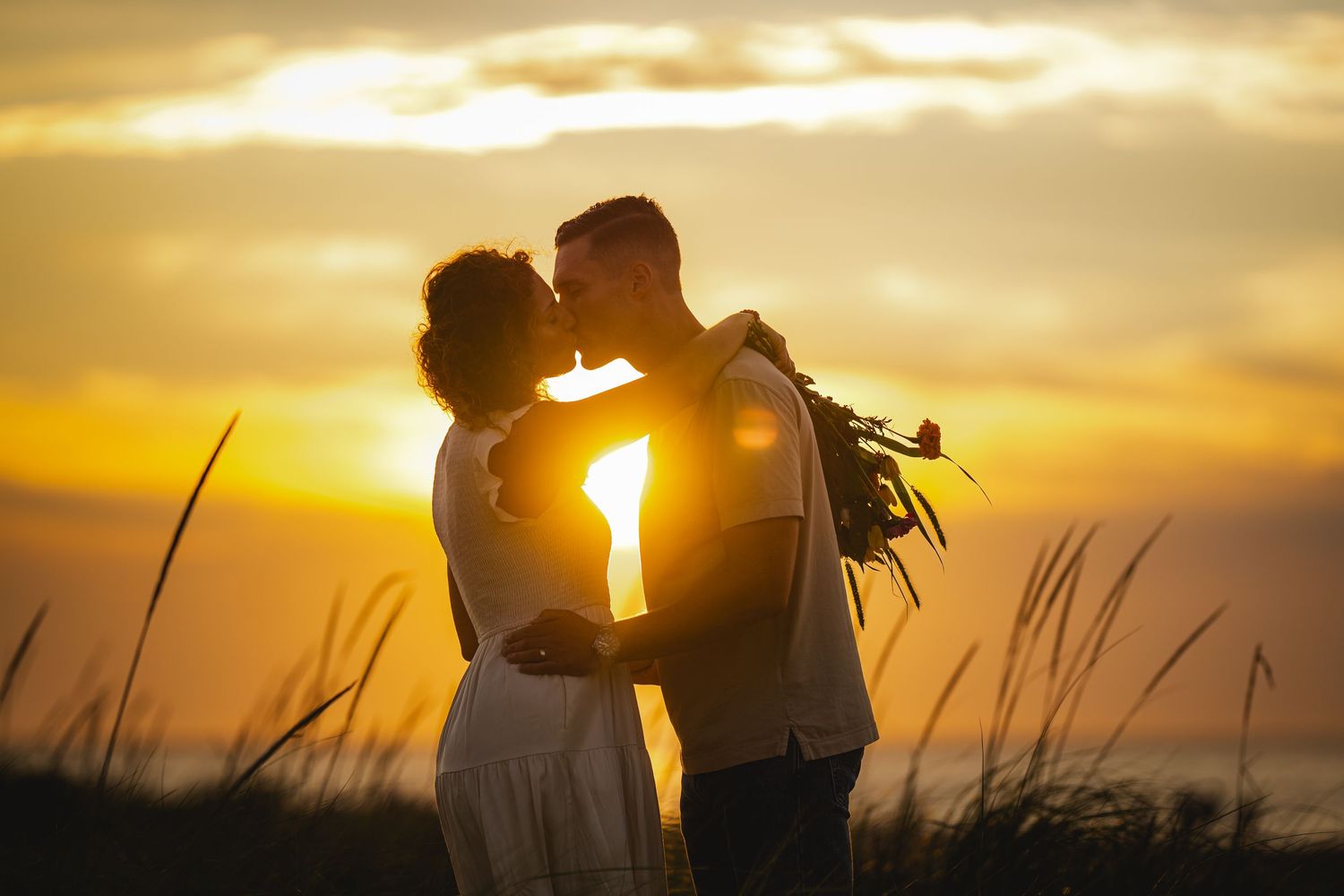 Silhouettes of a couple embracing on a beach at sunset with tall grass in the foreground.