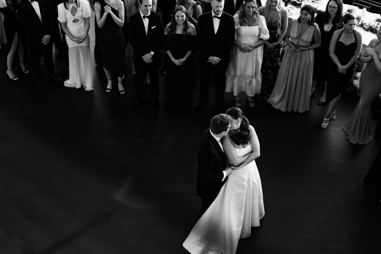 A black and white photo of a couple sharing their first dance at a wedding reception surrounded by guests.